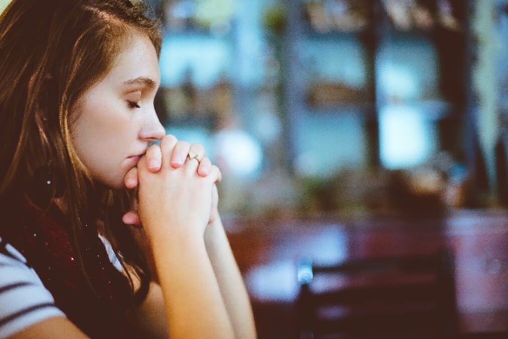 Uma mulher de aparência preocupada, com os olhos fechados e as mãos apoiadas no queixo, simbolizando a sensação de ansiedade e sobrecarga emocional.